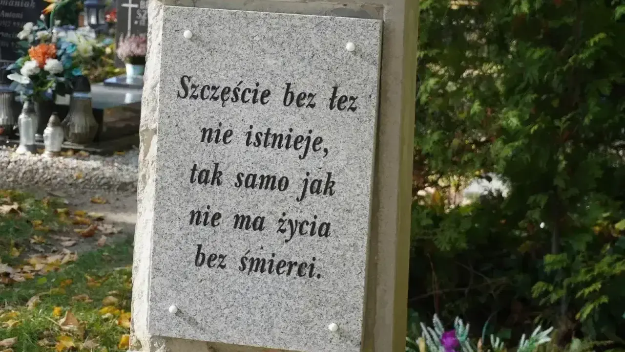 Kamienny nagrobek z napisem "Szczęście bez łez nie istnieje, tak samo jak nie ma życia bez śmierci". W tle widać inne groby i zieleń.