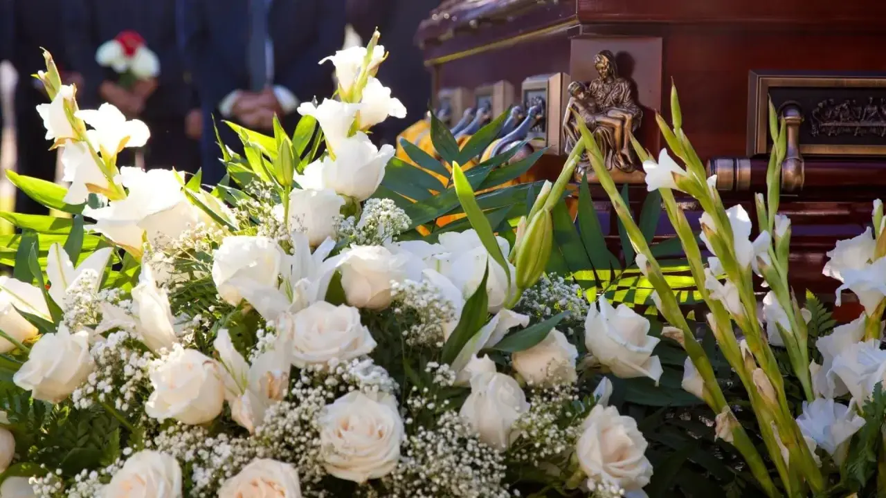 Elegancki wieniec pogrzebowy z białych róż i lilii, ozdobiony zielenią, przy trumnie. Cena 200 zł.