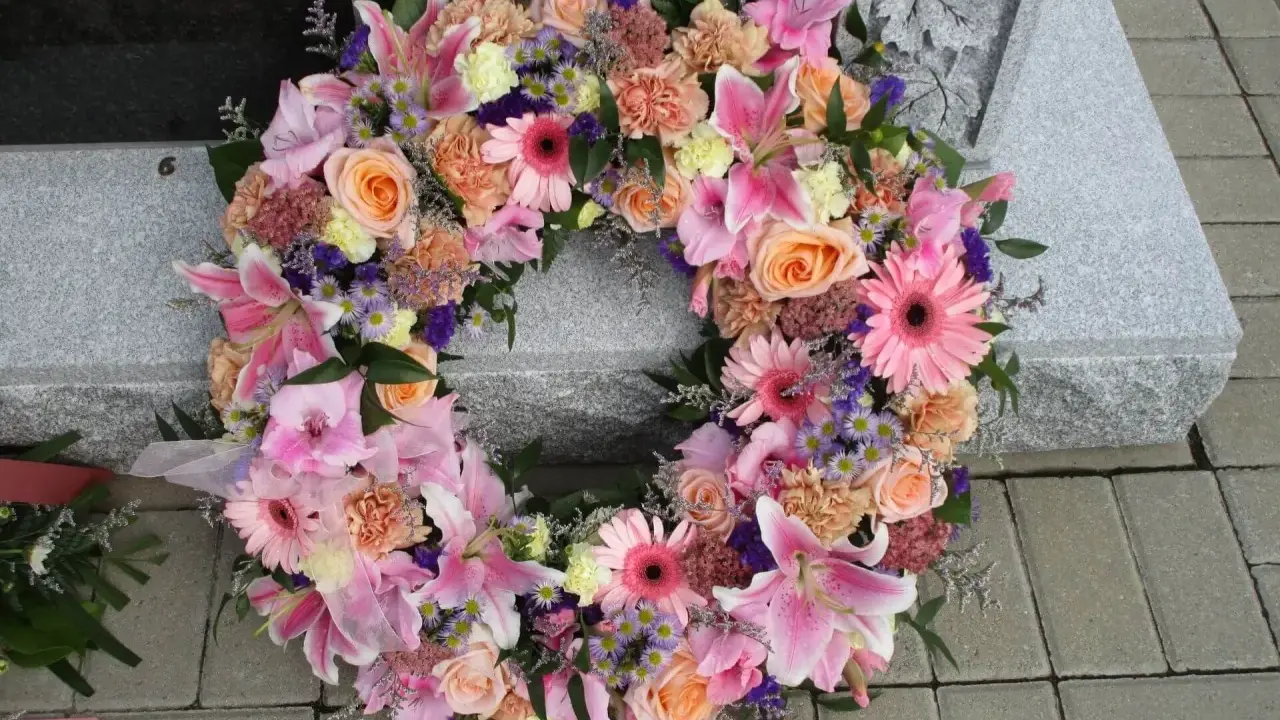 Piękna kompozycja funeralna z różowych lilii, róż i gerber, ozdobiona fioletowymi akcentami, spoczywa na kamiennej płycie nagrobnej.
