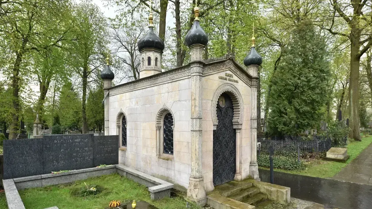 Elegancki, murowany grób z kopułami i ozdobnymi drzwiami, otoczony zielenią cmentarza.