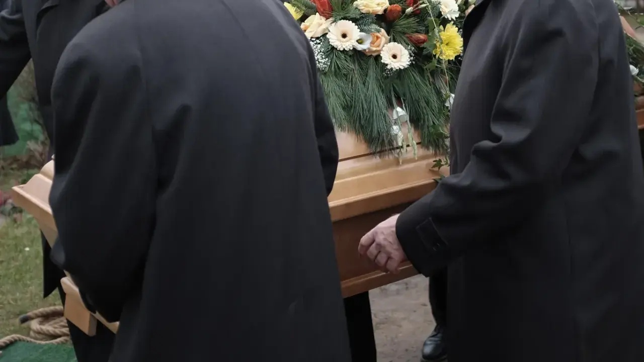 Mężczyźni w czarnych płaszczach niosą trumnę ozdobioną kwiatami. Kondukt pogrzebowy w żałobnej ciszy.