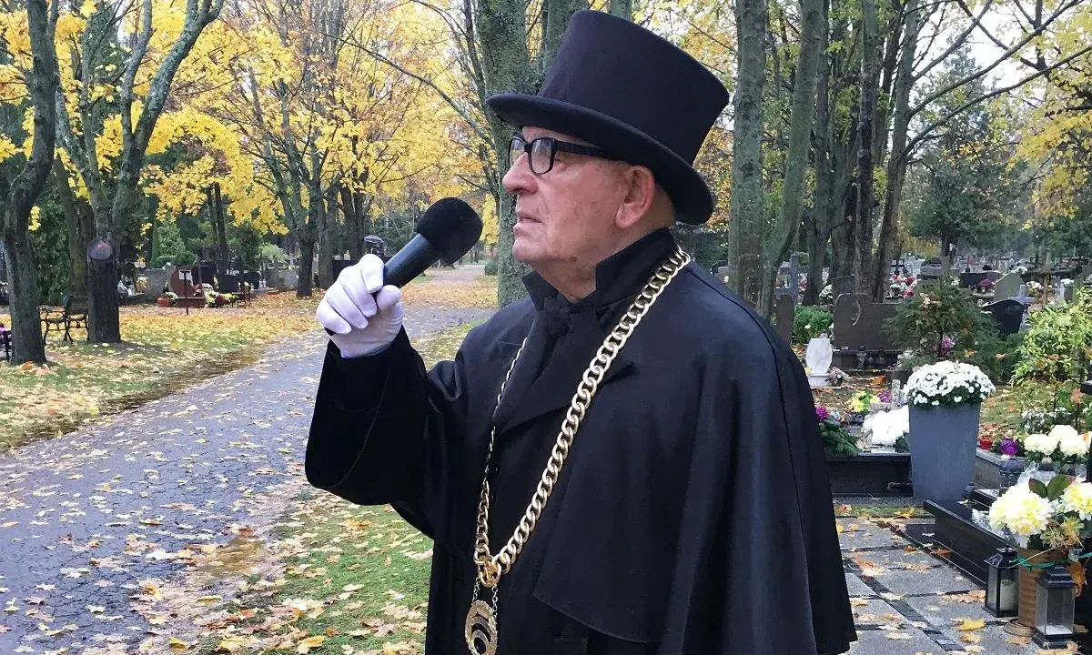 Uroczysty mistrz ceremonii pogrzebowej w czarnym stroju i cylindrze, z mikrofonem w ręku, przemawia na cmentarzu. Cena jego usług jest adekwatna do powagi chwili.