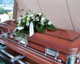 Brązowa trumna z wieńcem białych kwiatów. Na wstędze widnieje napis "GRANNY". Informacje, jak sprawdzić datę pogrzebu, można znaleźć w nekrologach.