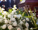 Elegancki wieniec pogrzebowy z białych róż i lilii, ozdobiony zielenią, przy trumnie. Cena 200 zł.