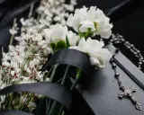 Białe kalie, drobne białe kwiatuszki i czarna wstęga na czarnym tle. Obok leży różaniec i krzyż.