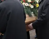Mężczyźni w czarnych płaszczach niosą trumnę ozdobioną kwiatami. Kondukt pogrzebowy w żałobnej ciszy.
