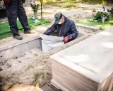 Ekshumacja zwłok po 40 latach. Pracownicy w masce i rękawiczkach wyciągają worek z grobu. Obok leży nowa trumna.