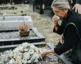 Kobieta w żałobie klęczy przy grobie, ocierając łzy. Bukiet kwiatów leży na ziemi. To smutne ostatnie pożegnanie.