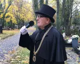 Uroczysty mistrz ceremonii pogrzebowej w czarnym stroju i cylindrze, z mikrofonem w ręku, przemawia na cmentarzu. Cena jego usług jest adekwatna do powagi chwili.