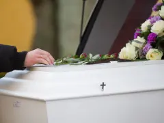 Dłoń z obrączką kładzie róże na białej trumnie, ozdobionej bukietem kwiatów i krzyżem.
