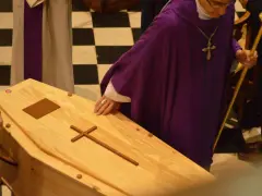 Uroczyste pożegnanie zmarłego w kaplicy. Ksiądz w purpurowej dalmatyce i z krzyżem na szyi błogosławi trumnę.