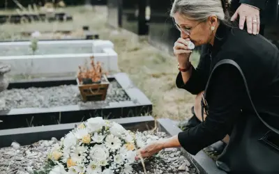 Kobieta w żałobie klęczy przy grobie, ocierając łzy. Czy zasiłek pogrzebowy należy się każdemu? Wieniec z białych i żółtych kwiatów leży na ziemi.