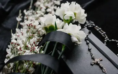 Białe kalie, drobne białe kwiatuszki i czarna wstęga na czarnym tle. Obok leży różaniec i krzyż.