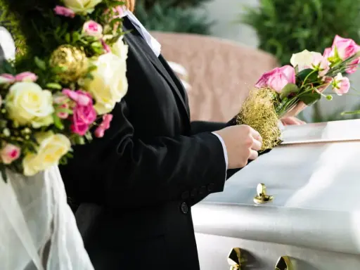 Osoba w czarnym garniturze kładzie bukiet róż na białej trumnie.