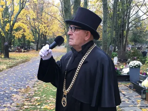 Uroczysty mistrz ceremonii pogrzebowej w czarnym stroju i cylindrze, z mikrofonem w ręku, przemawia na cmentarzu. Cena jego usług jest adekwatna do powagi chwili.