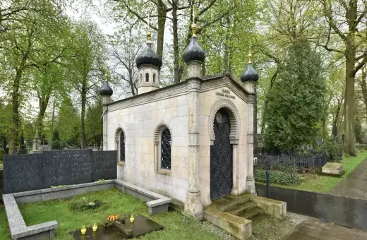 Elegancki, murowany grób z kopułami i ozdobnymi drzwiami, otoczony zielenią cmentarza.