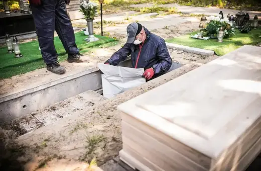 Pracownicy wykonują ekshumację, wyciągając biały worek z grobu. Obok leży drewniana trumna.