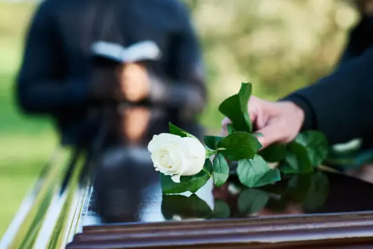 Biała róża na trumnie, w tle osoba czytająca z księgi. Chwila zadumy podczas wzruszającego kazania pogrzebowego.