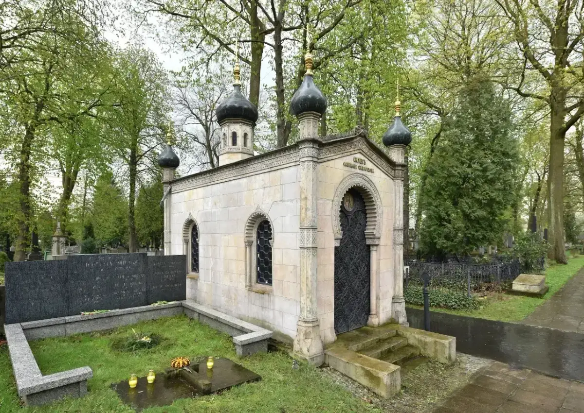 Elegancki, murowany grób z kopułami i ozdobnymi drzwiami, otoczony zielenią cmentarza.