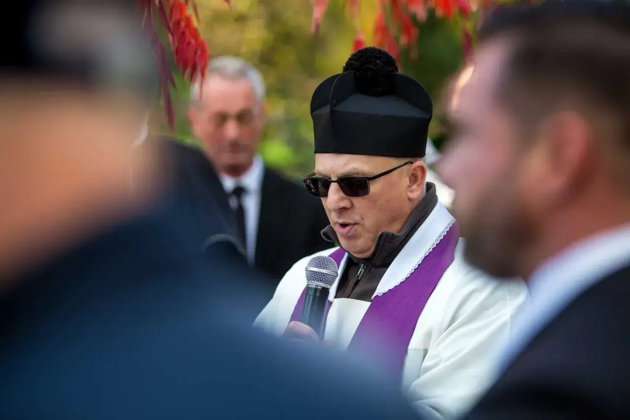Ksiądz w okularach i czarnej czapce z pomponem czyta pożegnanie zmarłego. W tle widać innych uczestników ceremonii.