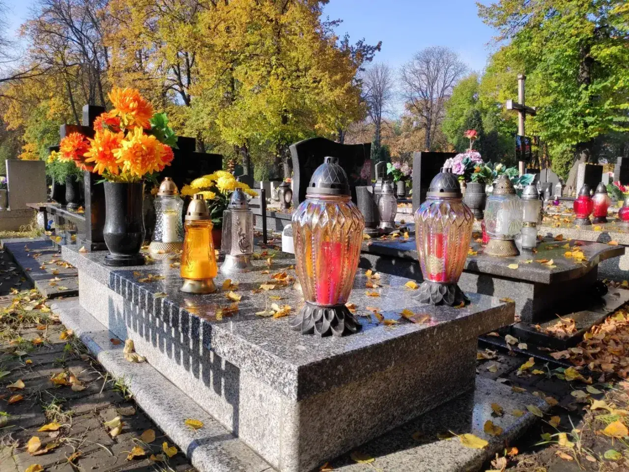 Znicze i kwiaty na kamiennym nagrobku. Jesienne liście pokrywają ziemny grób, tworząc melancholijną atmosferę.