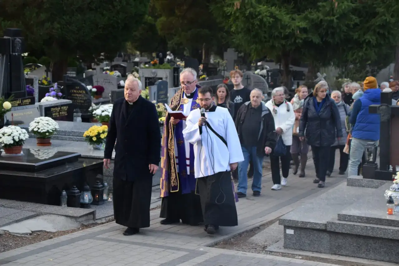Uroczysty kondukt pogrzebowy na cmentarzu. Ksiądz w stroju liturgicznym prowadzi żałobników wśród nagrobków i kwiatów.